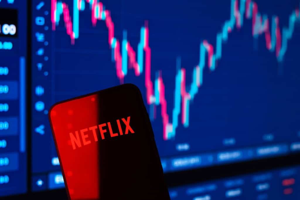Ao fundo, gráficos financeiros em uma tela; à frente, logo da Netflix em um smartphone