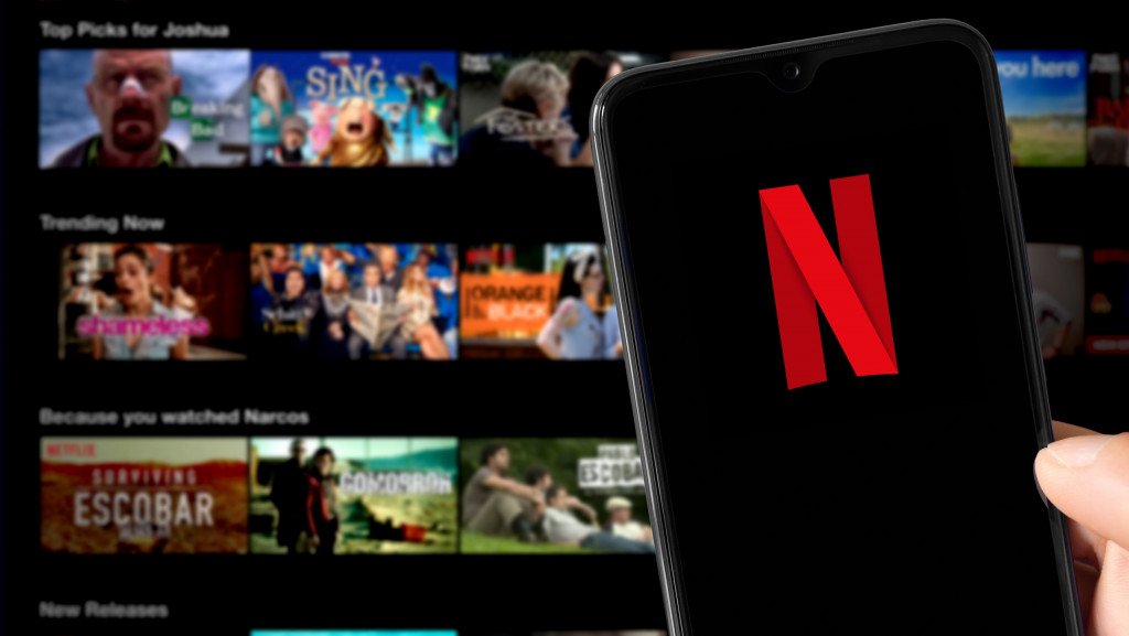 Ao fundo, desfocado, tela inicial da Netflix em uma TV; à frente, celular com o logo da empresa na tela